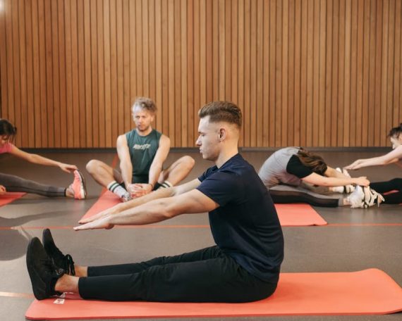 A diverse group of adults practicing yoga stretches on mats in a wooden studio.